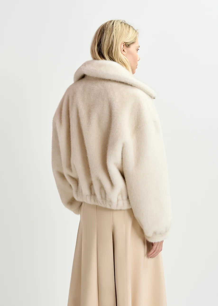 A woman with blonde hair is shown from the back wearing an oversized bomber silhouette with the white Genz Faux Fur Bomber Jacket by Essentiel Antwerp and a beige skirt against a plain background.