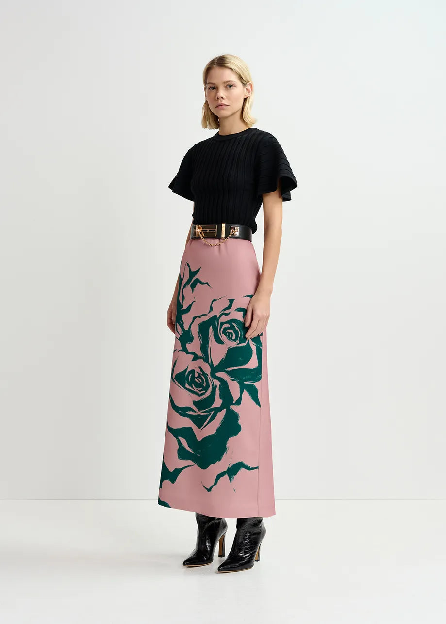 A woman standing against a plain background, wearing an Essentiel Antwerp Georgie rib-knitted top in black, a patterned pink skirt with green designs, a black belt, and black ankle boots.