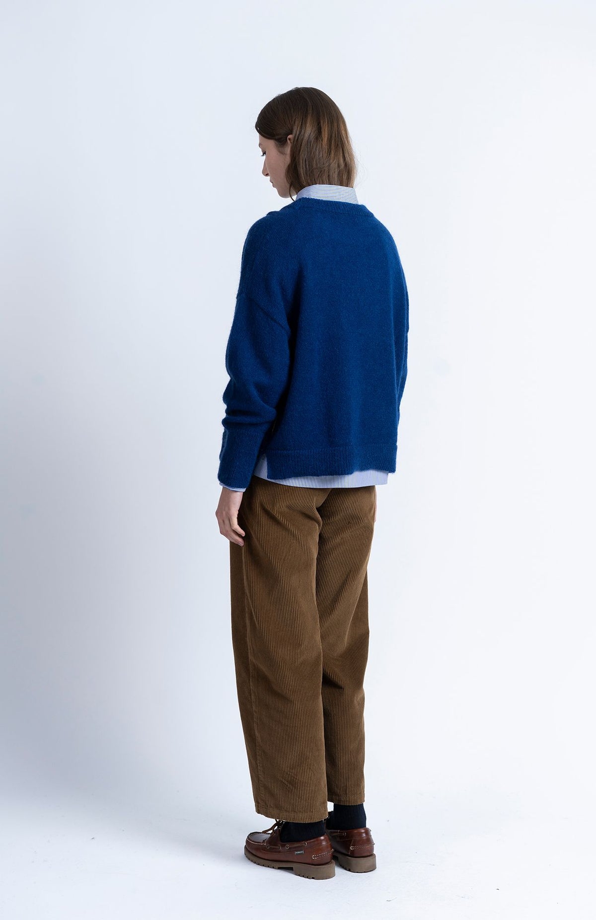 A person viewed from behind, wearing a navy Loreak Mendian Geroa Jumper over a striped button-down shirt, paired with brown corduroy trousers and brown leather boat shoes.