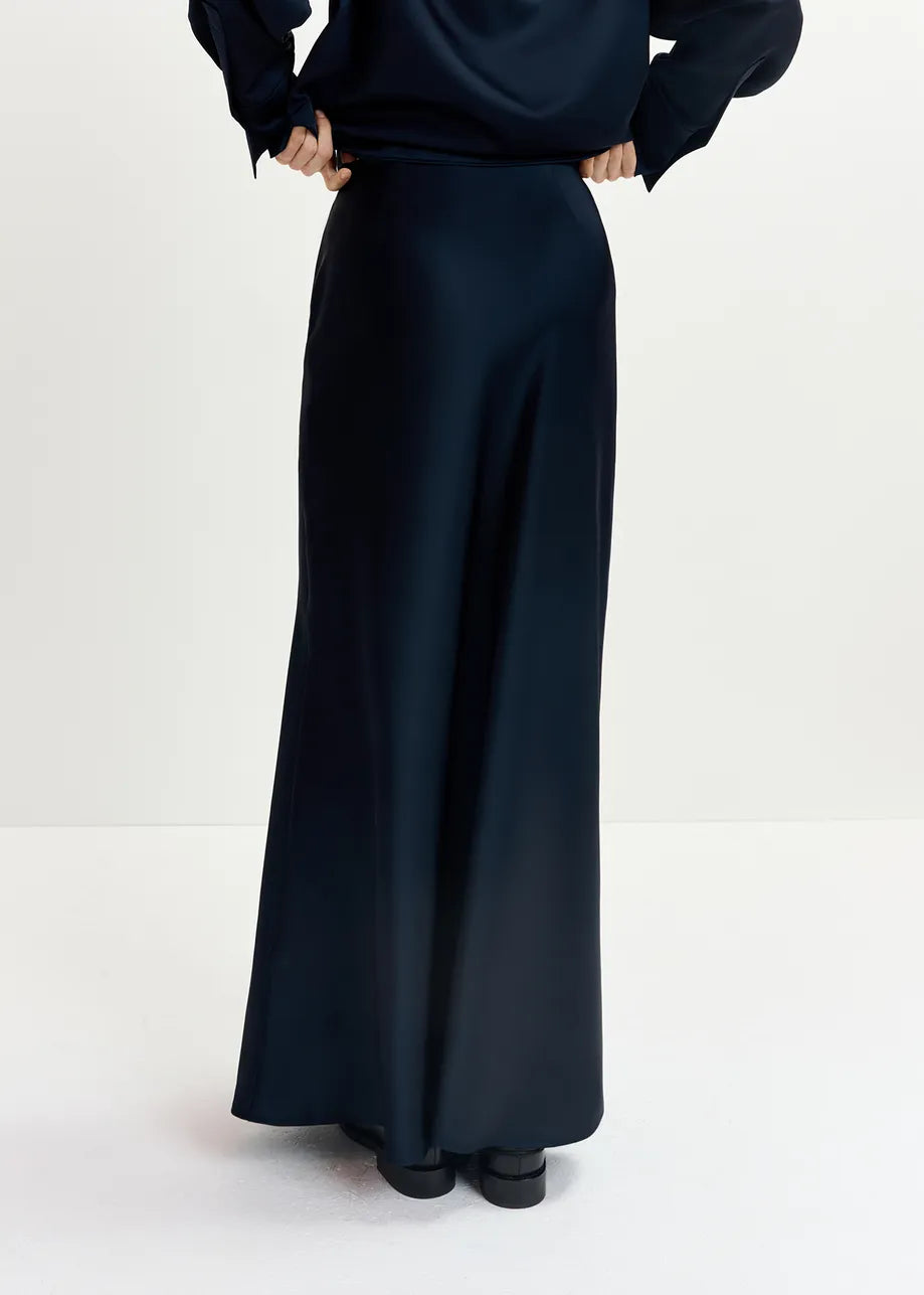 A person wearing a navy Giada Satin Maxi Skirt by Essentiel Antwerp, made from a recycled polyester-blend satin, and dark shoes stands with their back to the camera against a plain light-colored background.