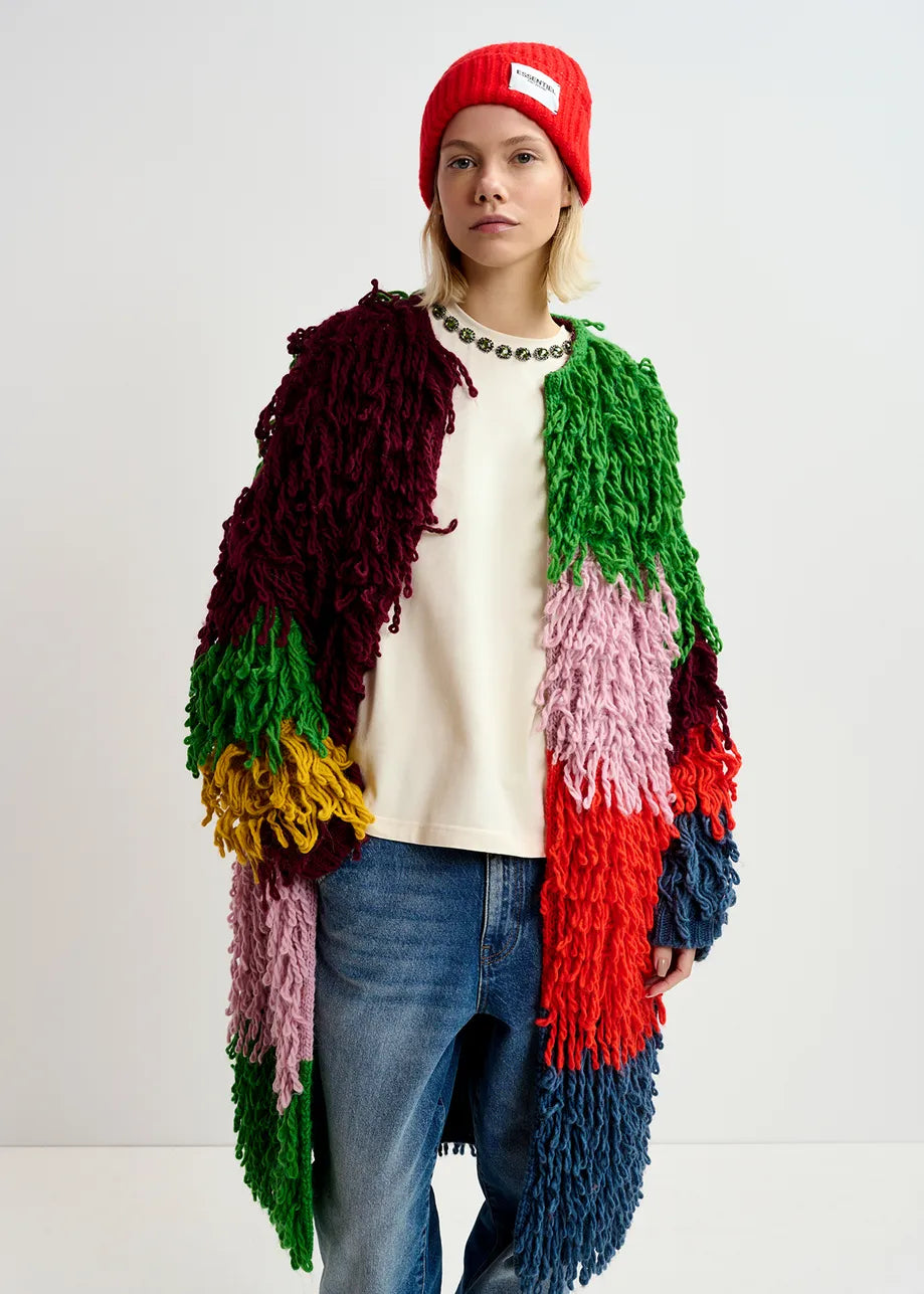 A person wearing the Gingle Oversized Knit - Multicolour from Essentiel Antwerp, paired with a white shirt, blue jeans, and a red beanie stands against a plain background. This oversized outerwear features unique loop-stitch knit details that add texture and flair to the versatile fit.