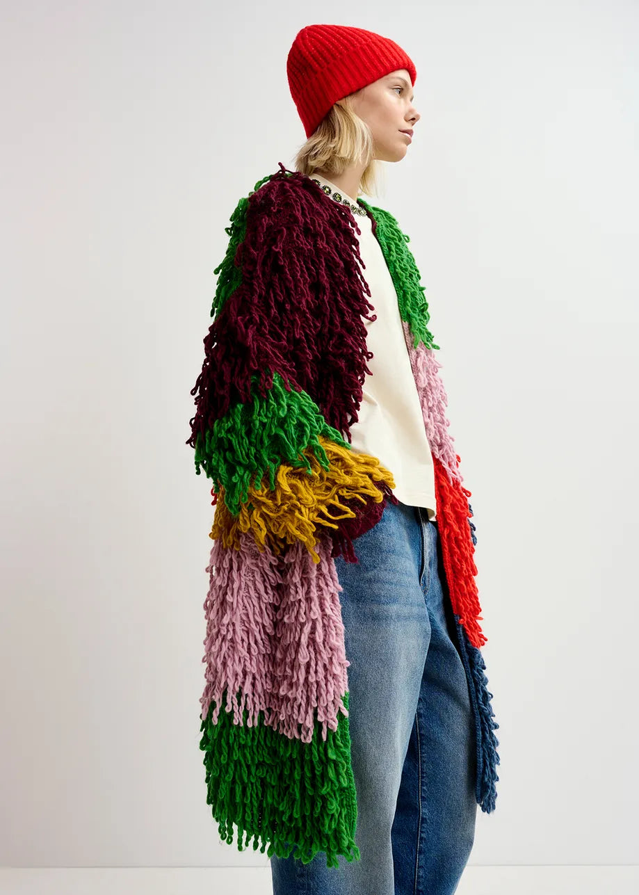 A person standing in profile is wearing the Essentiel Antwerp Gingle Oversized Knit in multicolor, paired with a red beanie, white shirt, and blue jeans against a plain background.