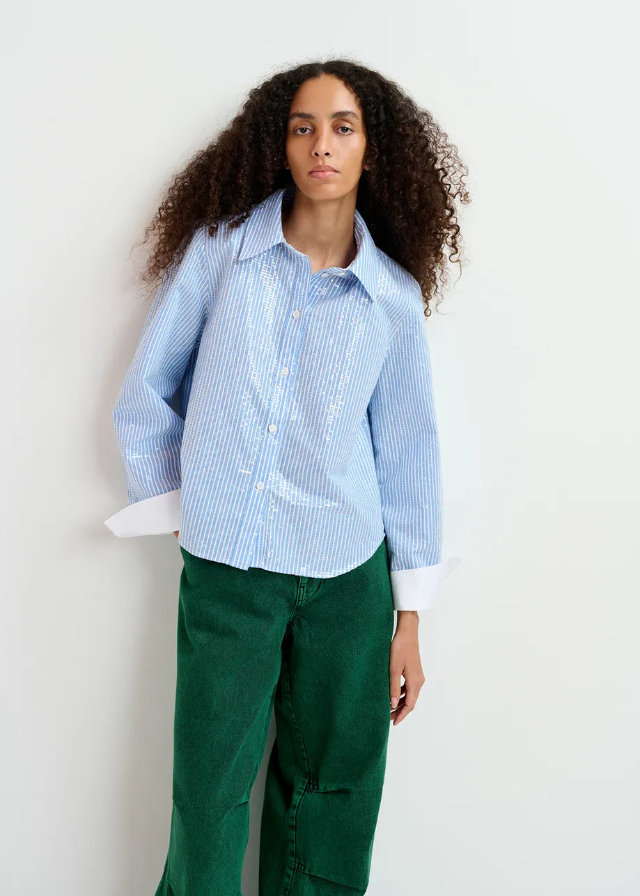 A person with long curly hair wears a blue Giuliano sequin-embellished shirt from Essentiel Antwerp and green pants, standing against a plain white background.