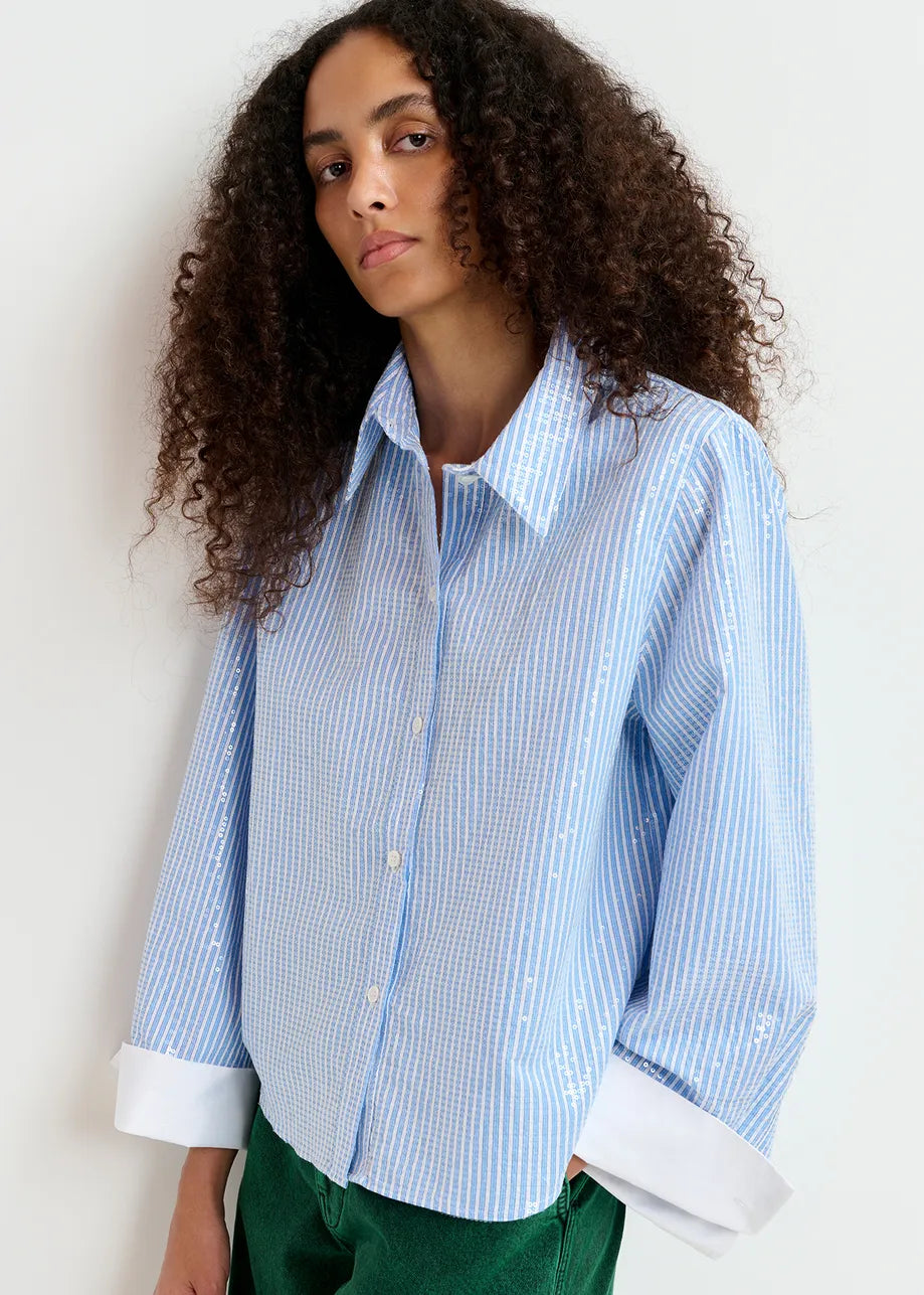 A person with curly hair wears the Giuliano Sequin-embellished Shirt in blue by Essentiel Antwerp, standing against a light background.