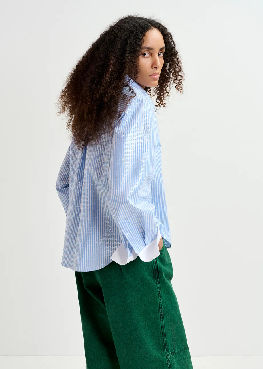 A person with long, curly hair wearing the Essentiel Antwerp Giuliano Sequin-Embellished Shirt in blue and green pants, turning slightly to the side while looking at the camera. The background is a plain, light-colored wall.