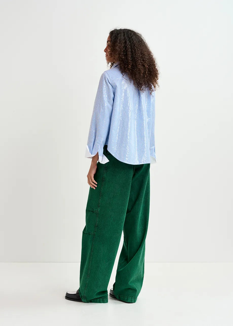 A person with curly hair stands facing away, wearing a light blue Giuliano Sequin-embellished Shirt by Essentiel Antwerp and green, loose-fitting pants. The background is plain white.