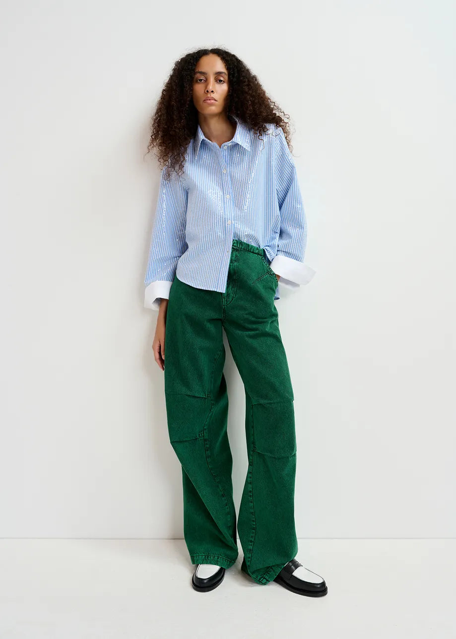 A woman with curly hair wears an Essentiel Antwerp Giuliano sequin-embellished shirt in blue and green wide-leg pants. She stands against a plain white background.