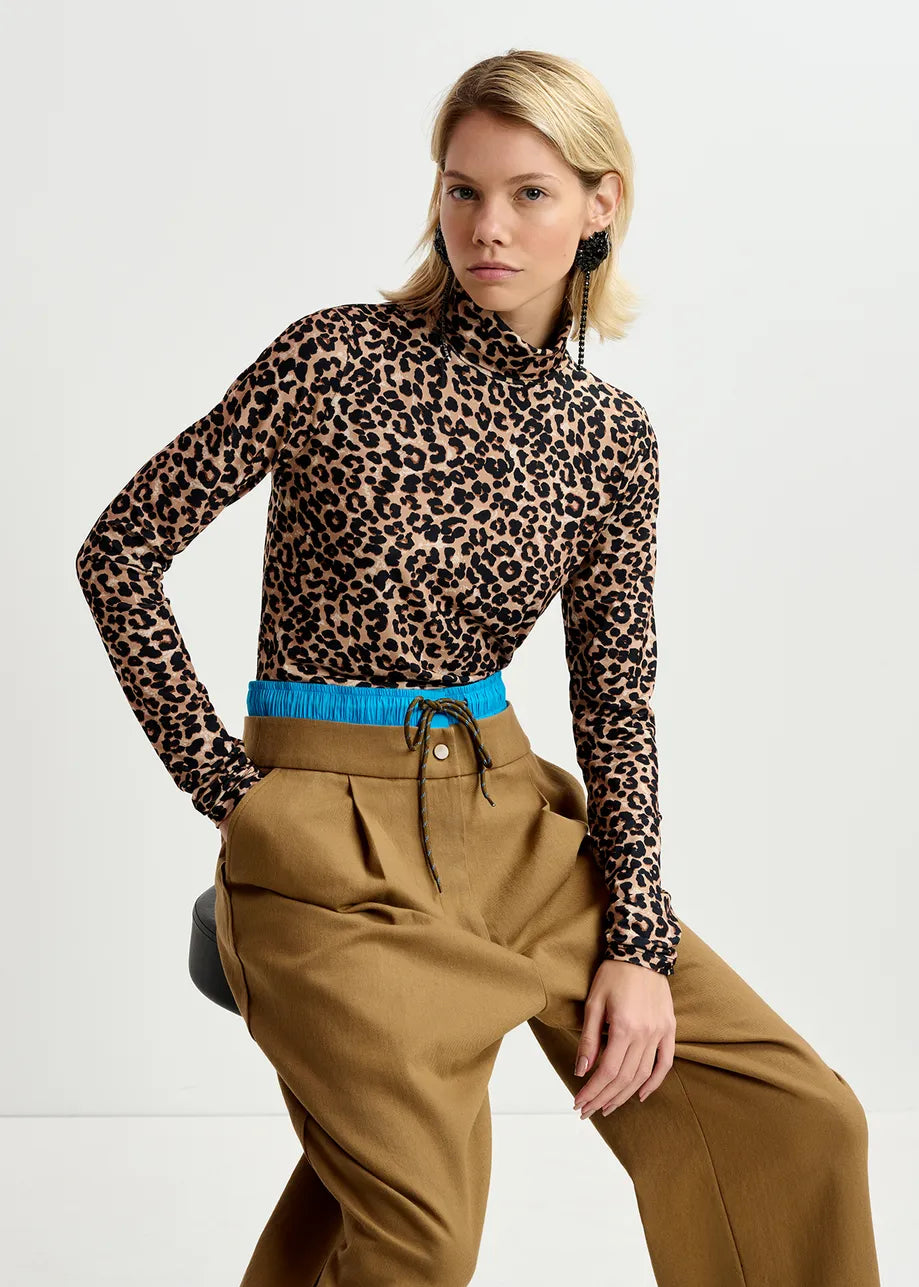 A person wearing the Essentiel Antwerp Going Turtleneck Top in leopard print, beige high-waisted pants with a blue waistband, and black earrings, posed in a sitting position.