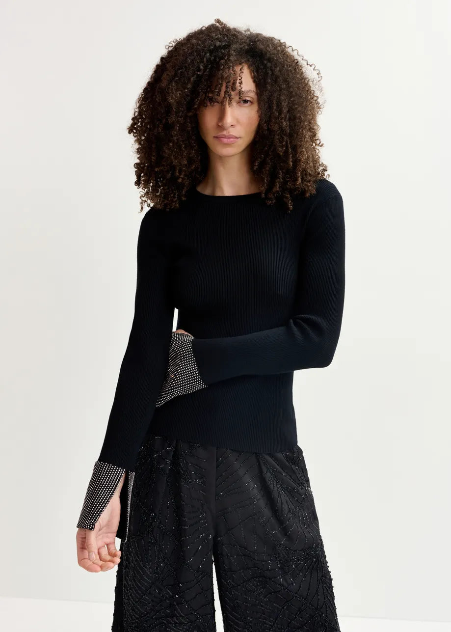 A person with curly hair stands against a plain background, wearing the Essentiel Antwerp Gomi Rib-knit Sweater in black, which features figure-hugging design and rhinestone-embellished cuffs, paired with black patterned pants.