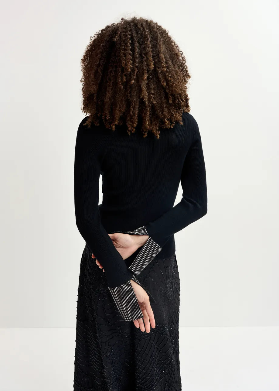 A person with curly hair is seen from behind, wearing the Essentiel Antwerp Gomi Rib-knit Sweater - Black and a textured skirt. Their arms are crossed behind their back, showcasing the rhinestone-embellished cuffs of the sweater that subtly shimmer.