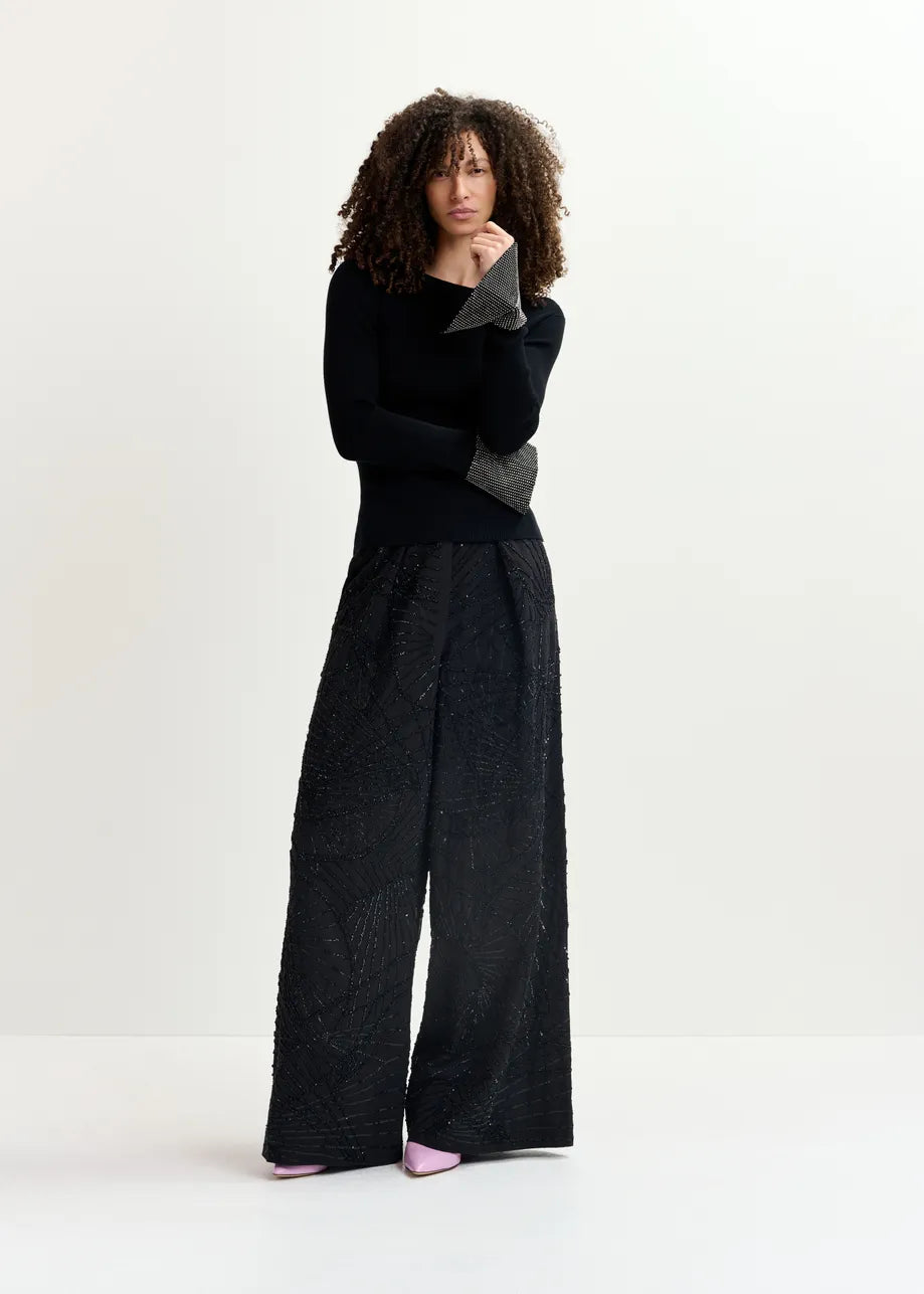 A person with curly hair stands against a plain background, wearing the Gomi Rib-knit Sweater from Essentiel Antwerp and wide-leg black pants.