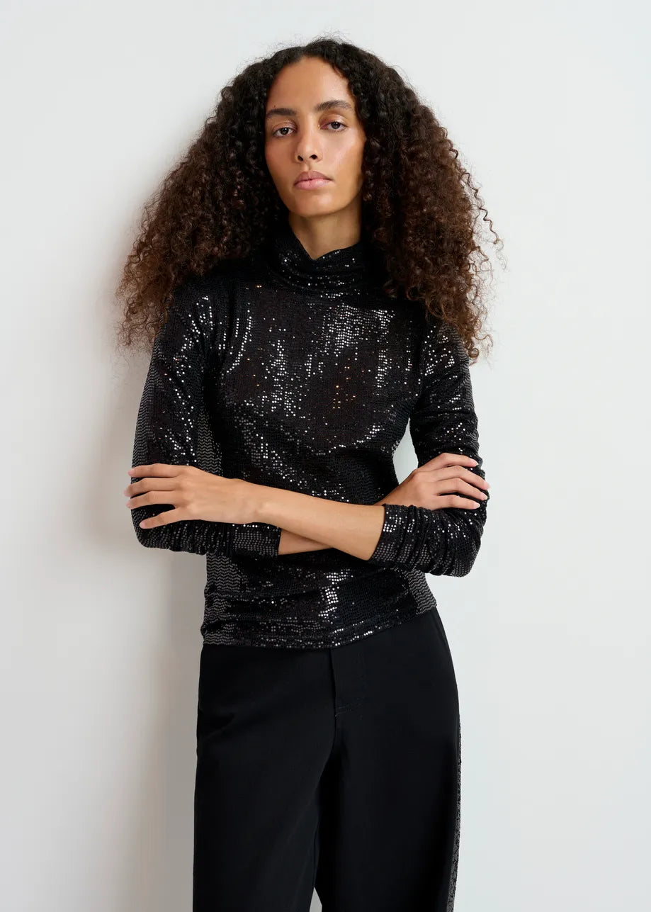 A person with curly hair is wearing the Gong Turtleneck Top in black from Essentiel Antwerp, featuring a glittery finish, and is standing against a plain white background.