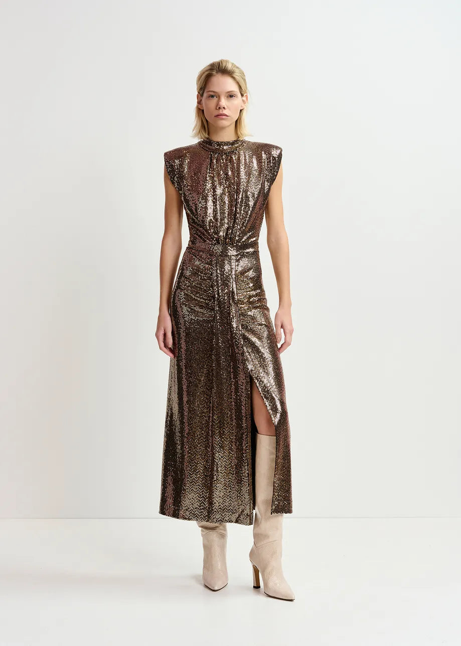 Wearing the Gonnak Dress - Gold by Essentiel Antwerp, which flaunts a stunning sequined metallic design, a figure-hugging fit, and a front slit, the individual pairs it with beige knee-high boots and poses against a plain white backdrop.