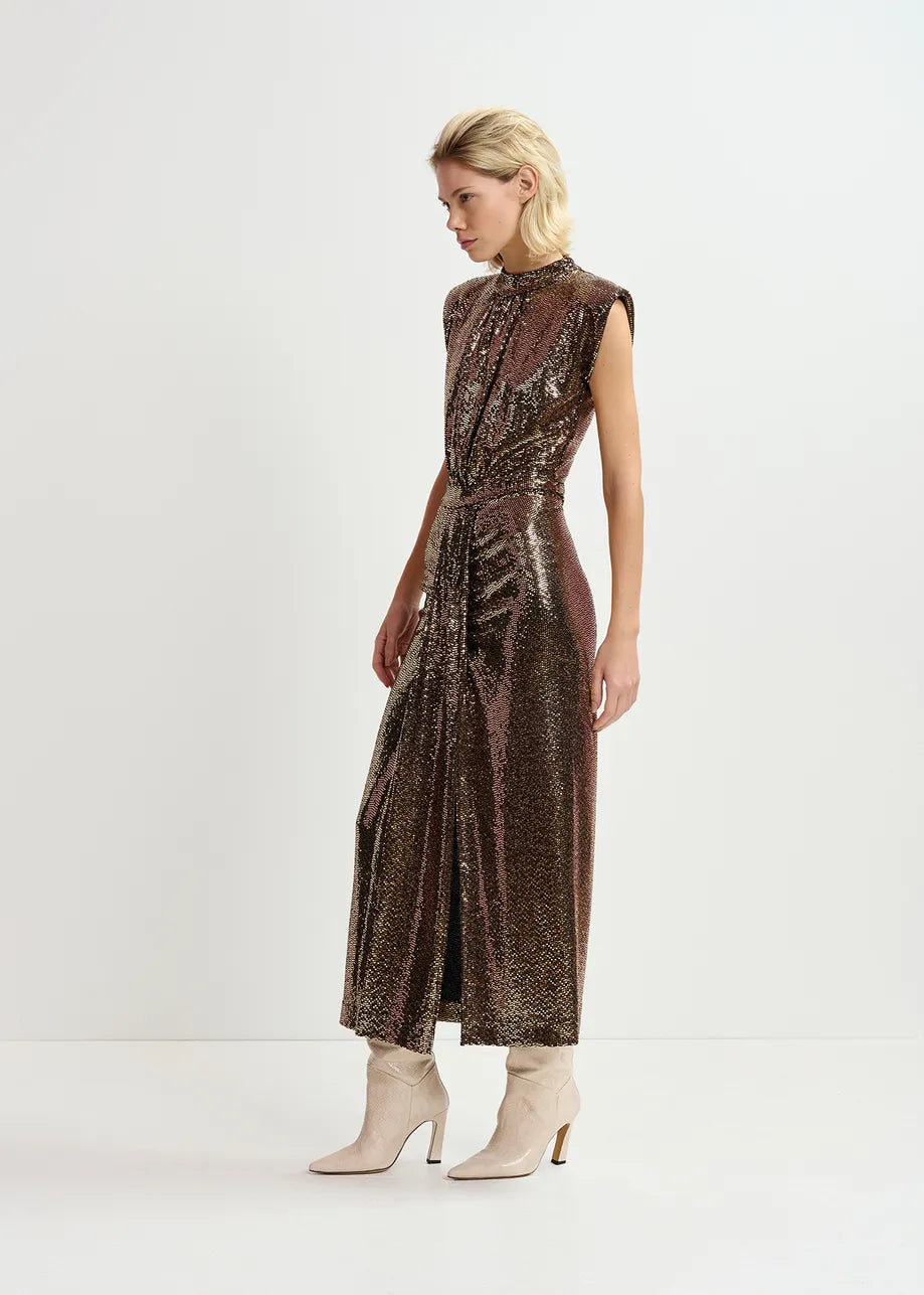 A person poses in an Essentiel Antwerp Gonnak Dress - Gold, featuring a sleeveless design with metallic sequins crafted from stretch-jersey fabric. The form-fitting silhouette is stylishly paired with light-colored ankle boots against a plain white backdrop.