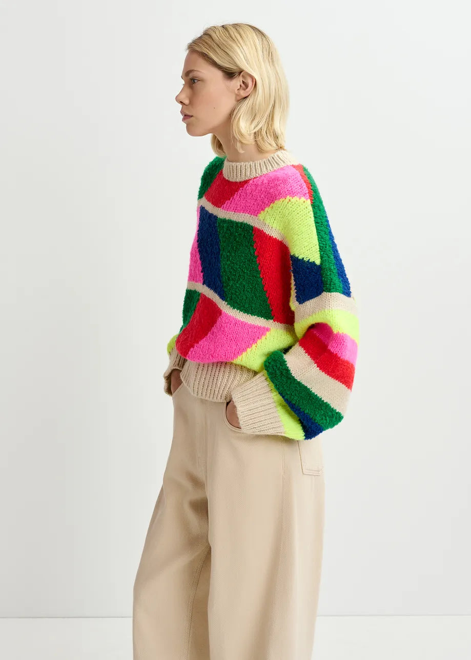 A person with blonde hair is standing sideways wearing a vibrant, multi-colored, geometric Gradation Knit Sweater by Essentiel Antwerp and light beige pants.