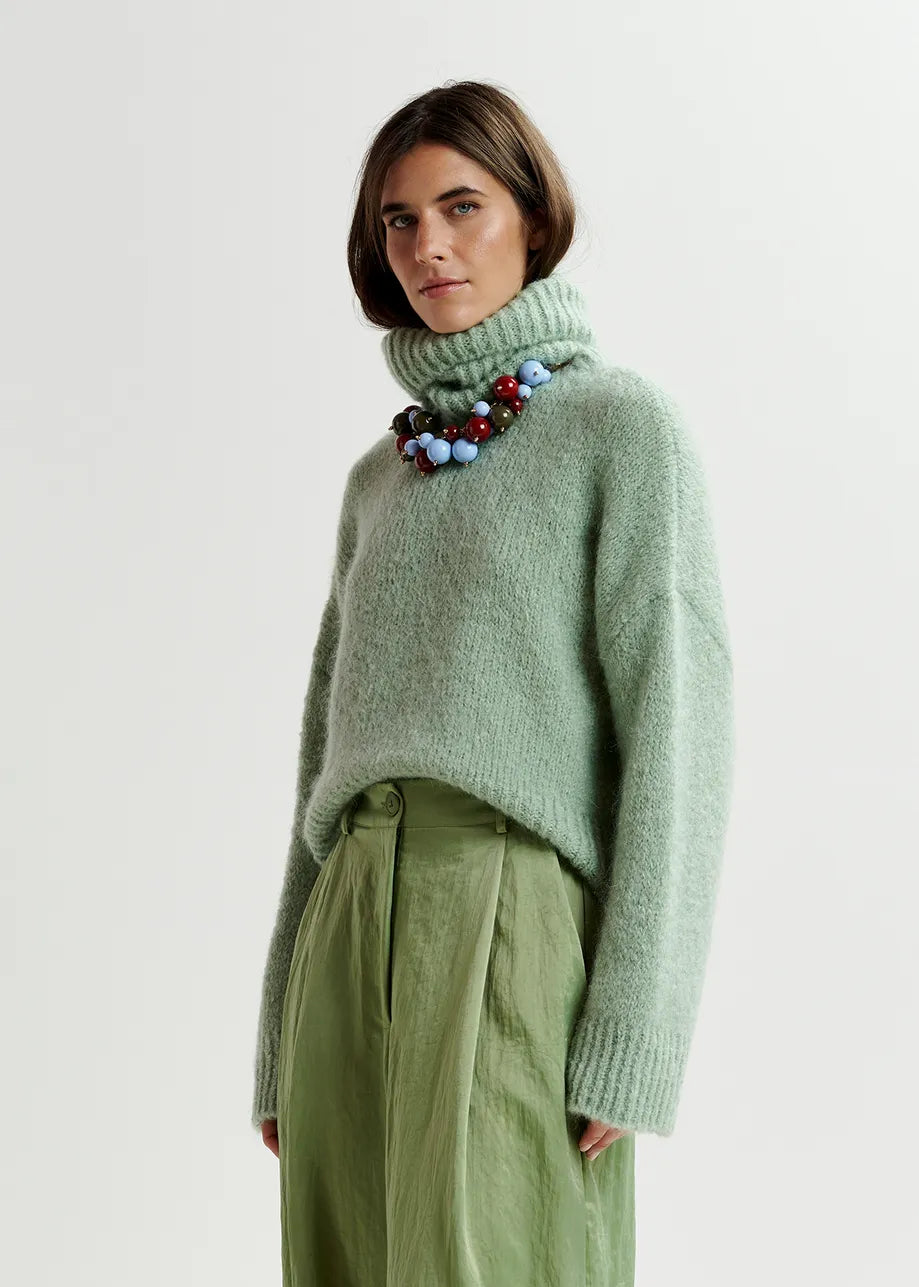 A person is wearing the Essentiel Antwerp Griffin Knit Turtleneck in light green, olive-green pleated pants, and a colorful beaded necklace, standing against a plain background.