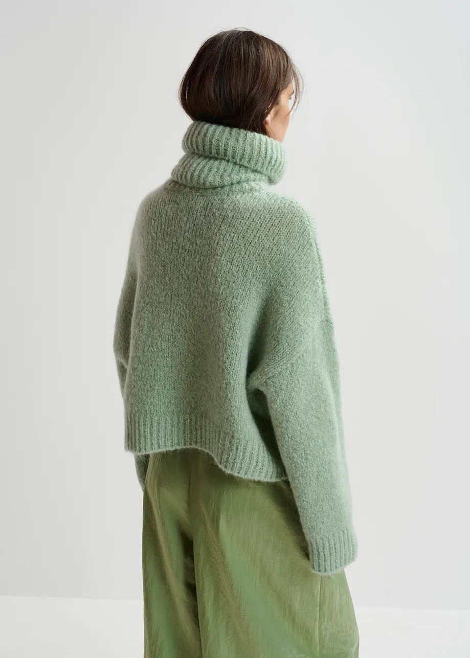 A person with brown hair, shown from the back against a plain background, is wearing the Griffin Knit Turtleneck in light green by Essentiel Antwerp along with matching green pants, featuring an oversized silhouette.