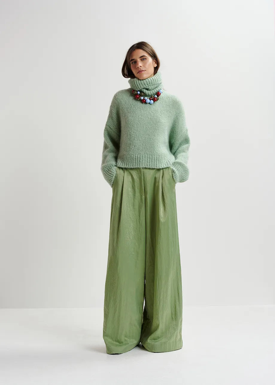 A person stands against a plain background wearing the Essentiel Antwerp Griffin Knit Turtleneck in light green, wide-legged green pants, and a colorful beaded necklace.