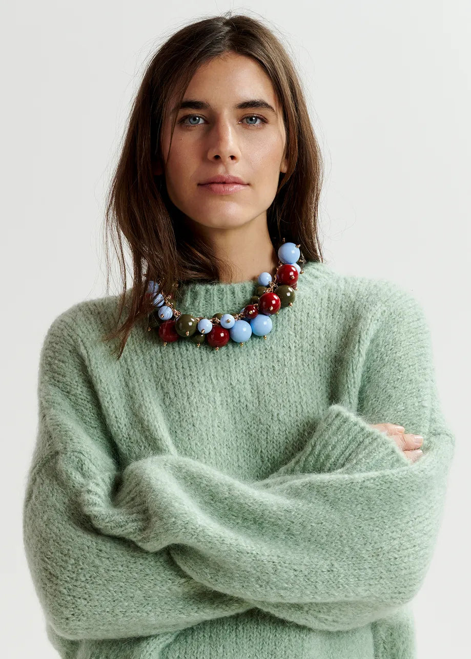 A person with long hair wears a light green Essentiel Antwerp Griffin Knit Turtleneck made from a mohair-blend and a colorful beaded necklace, standing with arms crossed.