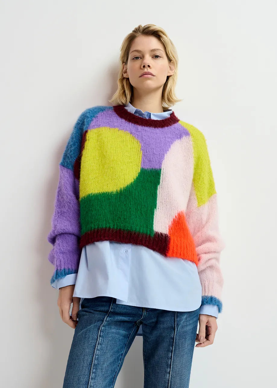 A person with short blonde hair stands against a plain white background, wearing Essentiel Antwerp's Groupie Knit Sweater in Multicolour over a light blue shirt, paired with blue jeans.