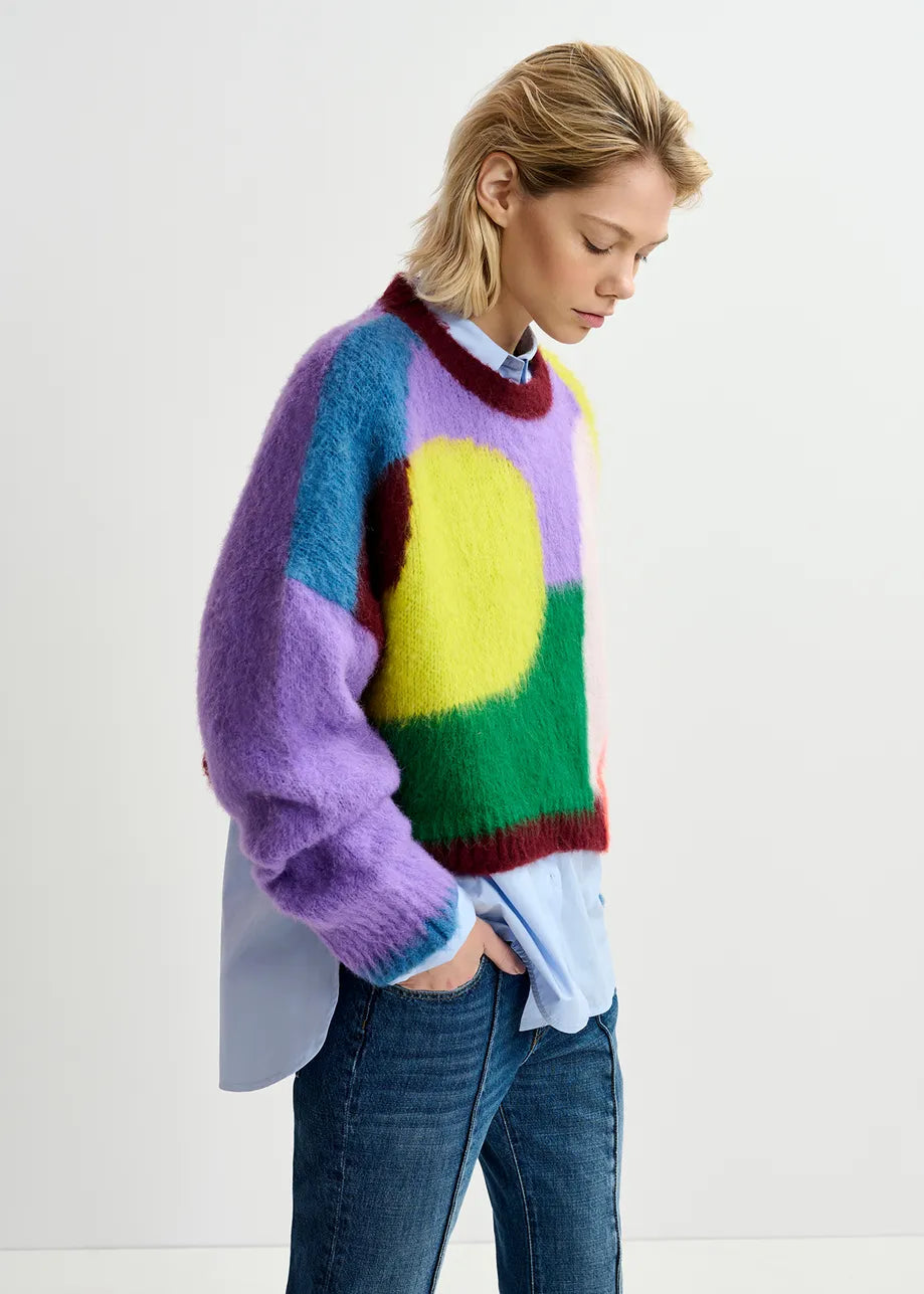 A person with short hair, standing against a plain background, is wearing an Essentiel Antwerp Groupie Knit Sweater - Multicolour over a light blue shirt and blue jeans, their head slightly tilted downwards.