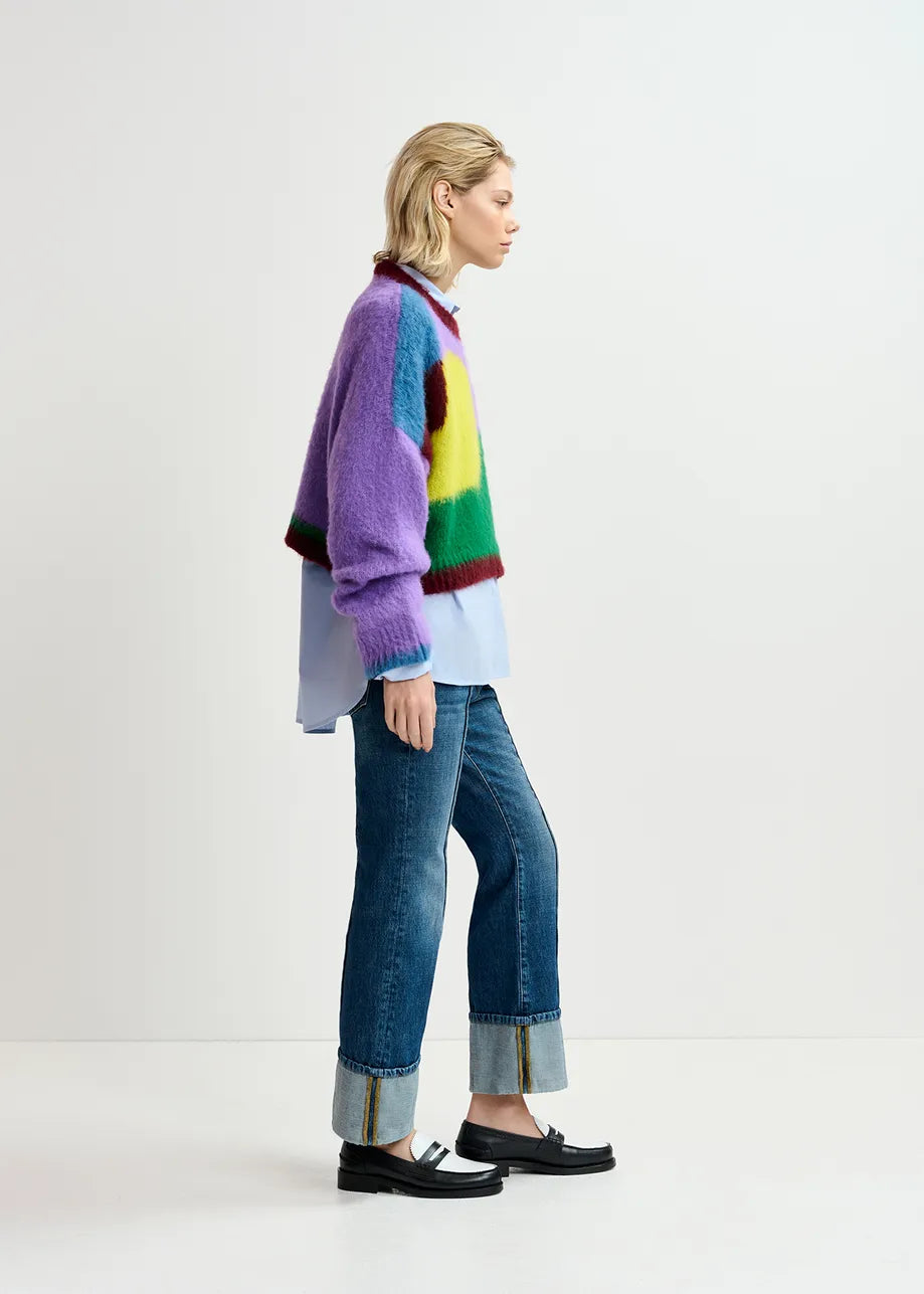 A person standing sideways wearing the Essentiel Antwerp Groupie Knit Sweater in Multicolour, paired with blue jeans cuffed at the ankles and loafers. The abstract intarsia-knitted design of the oversized silhouette sweater stands out as they gaze forward against a plain white background.