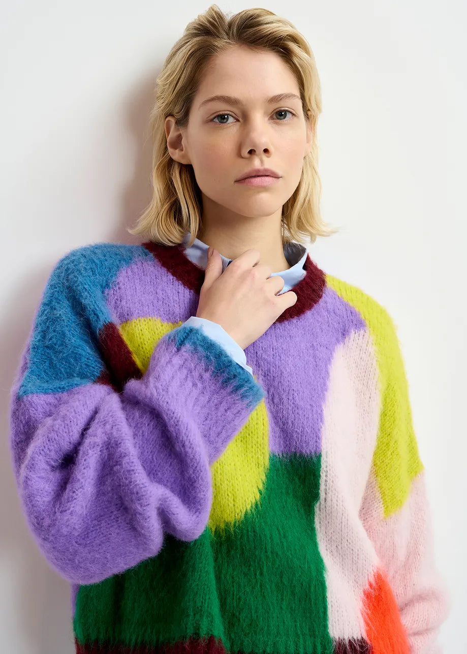 A person with short blonde hair wearing an oversized silhouette, multicolour Groupie Knit Sweater by Essentiel Antwerp stands against a plain backdrop, looking directly at the camera.