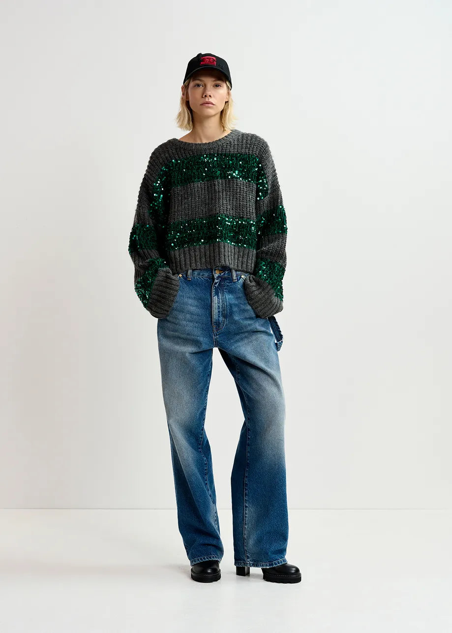 A person stands against a white background wearing the Group Sequin Knit - Green & Grey, an oversized silhouette sweater by Essentiel Antwerp, paired with blue jeans, black shoes, and a black cap.