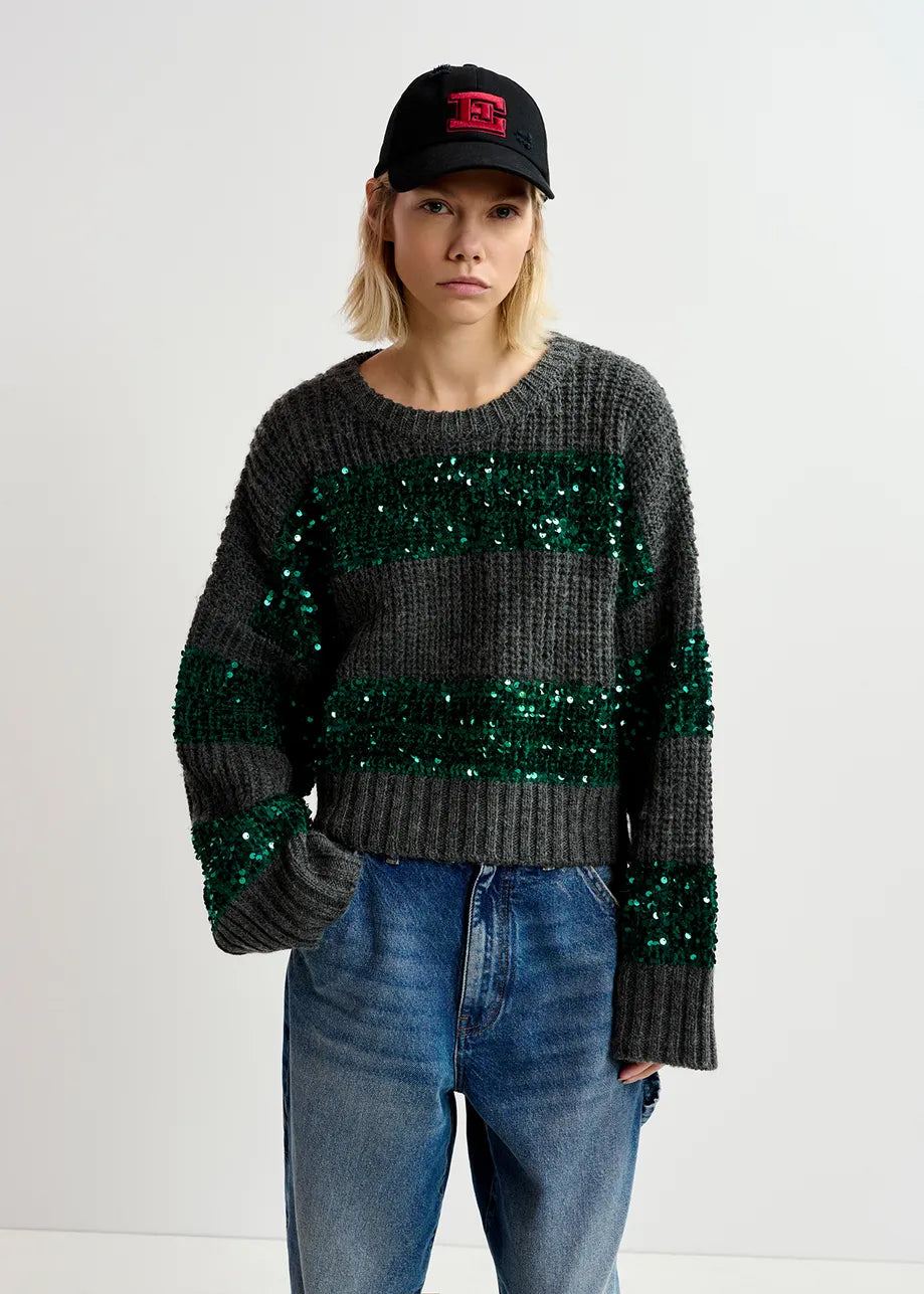 A person wearing the Group Sequin Knit - Green & Grey by Essentiel Antwerp, blue jeans, and a black baseball cap stands against a plain white background.