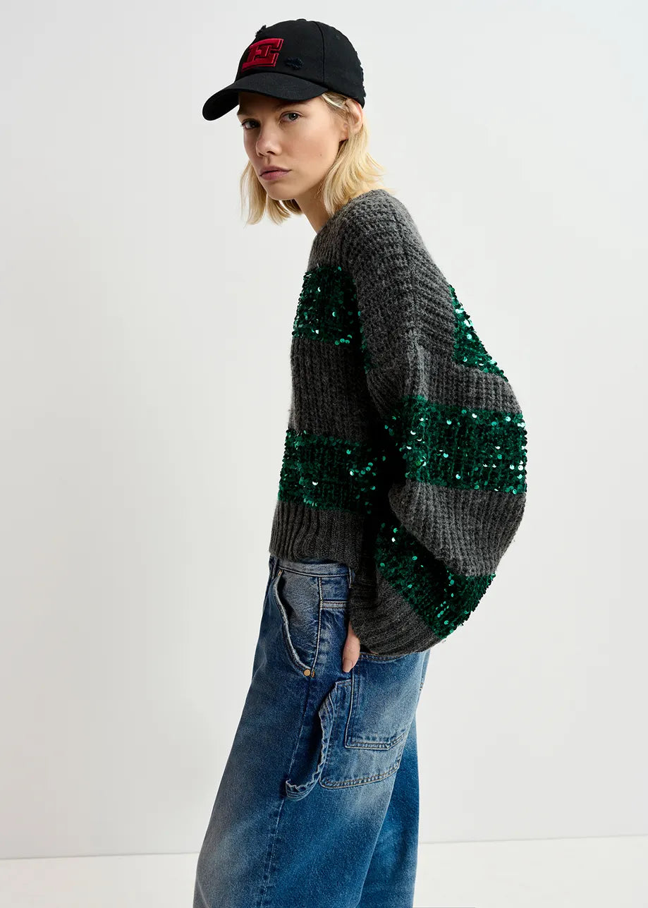 A person wearing the Essentiel Antwerp Group Sequin Knit - Green & Grey, paired with loose jeans and a black cap, is standing in profile against a plain background.