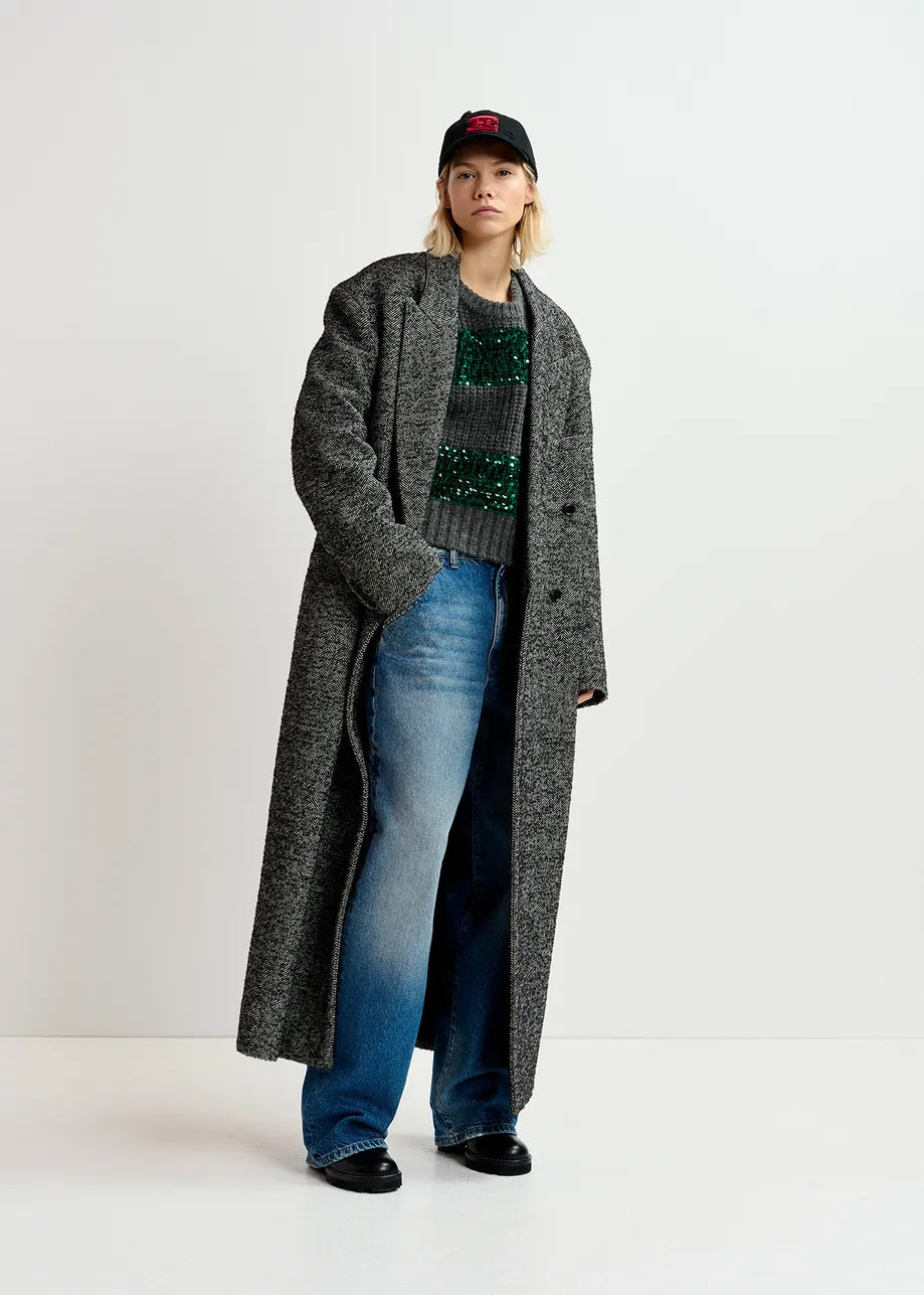 A person stands against a plain white background wearing a long gray coat with two-toned panels, baggy jeans, a chunky knit sweater called the Group Sequin Knit in green and grey by Essentiel Antwerp, black shoes, and a black baseball cap.