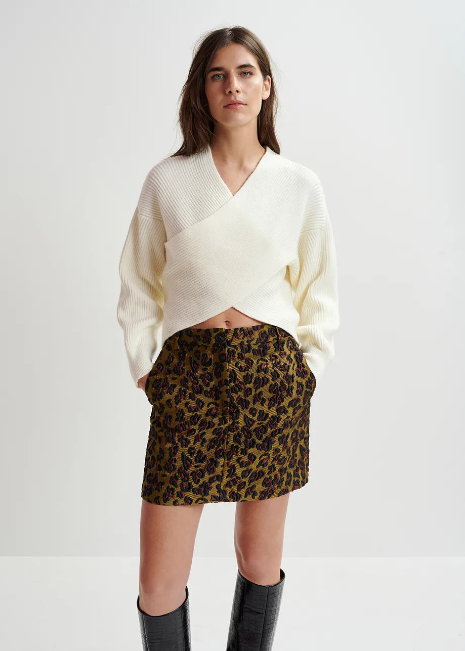 A woman is standing against a plain white background, wearing the Essentiel Antwerp Gustaph Cross-over Knit in white, a leopard print skirt, and black knee-high boots. She has her hands in her pockets and is looking forward.