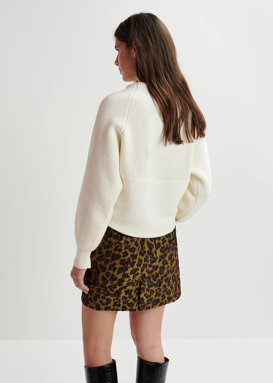 A person with long hair faces away from the camera, wearing the relaxed fit "Gustaph Cross-over Knit - White" by Essentiel Antwerp, paired with a leopard print skirt and black boots, standing against a plain white background.