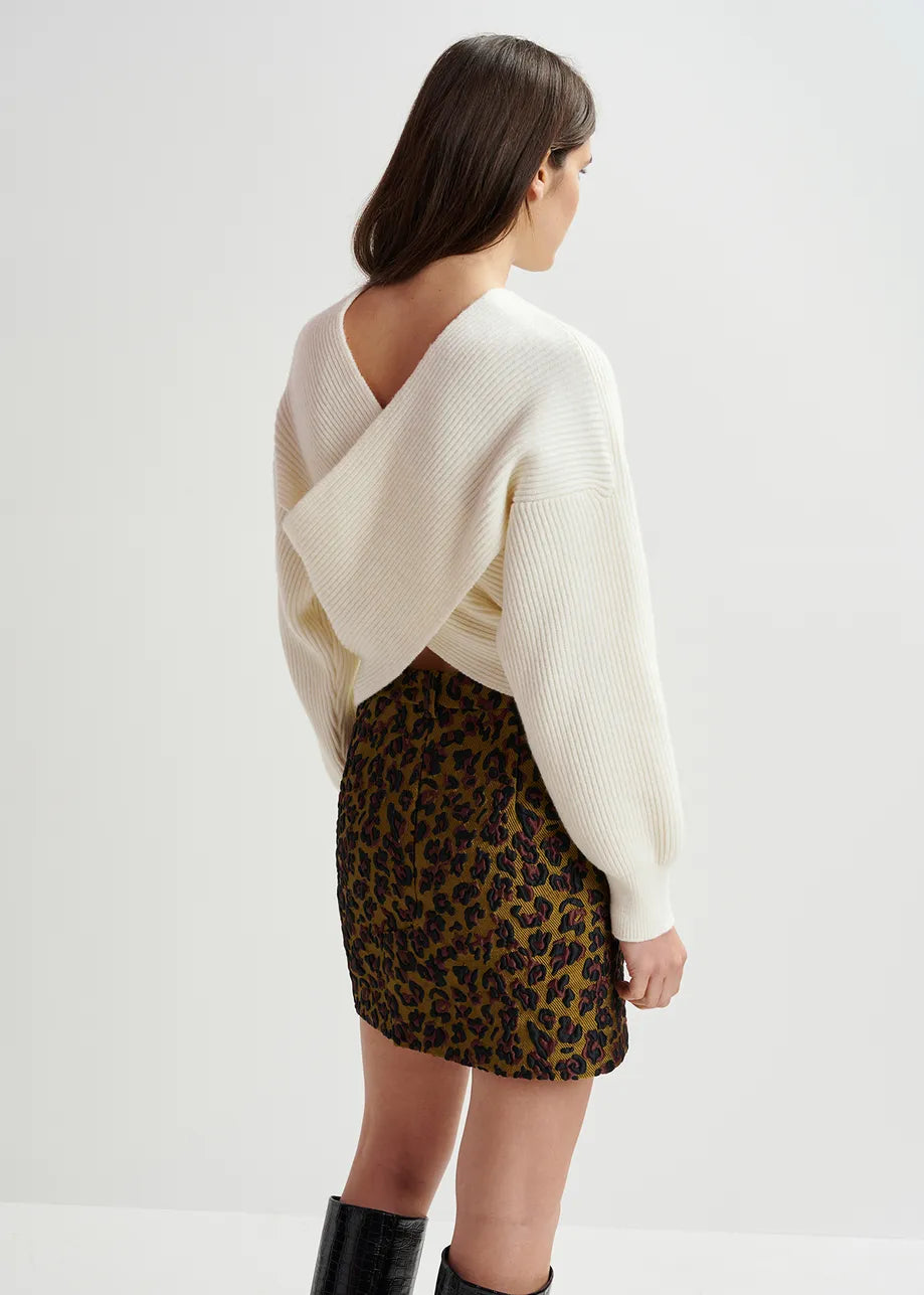 A person with long brown hair faces away, wearing the Essentiel Antwerp Gustaph Cross-over Knit in white, styled with a leopard print skirt and black boots.