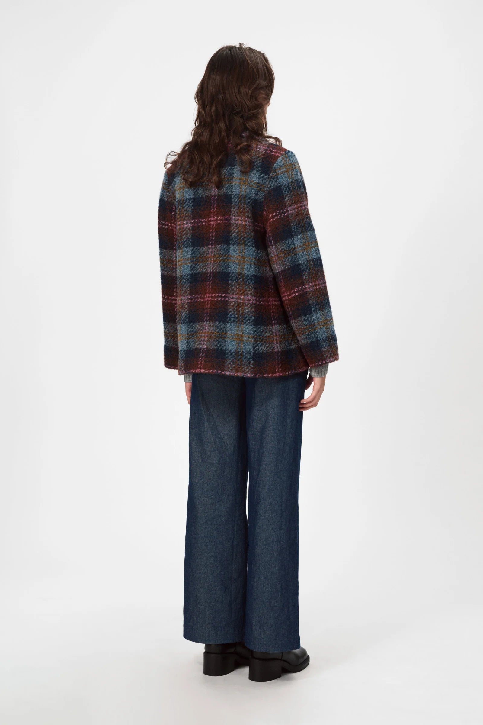 Seen from behind against a white background, a person with long brown hair wears a Nué Notes Glasgow Jacket - Fall Multi Check in an oversized fit, paired with wide-leg blue jeans and black shoes.
