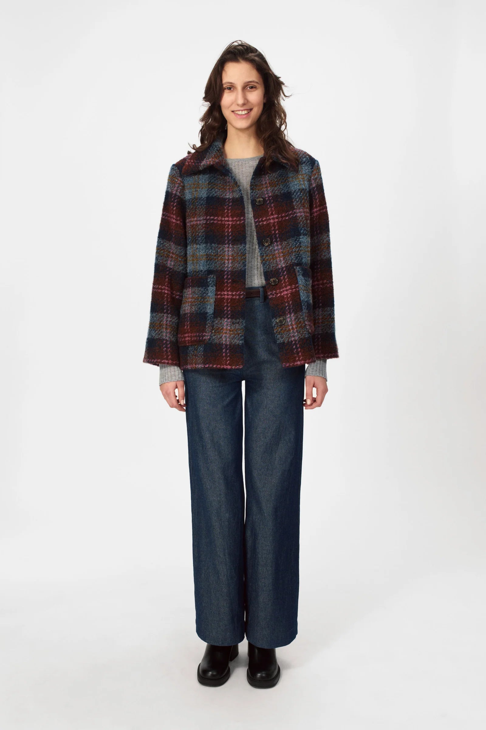 A woman stands against a white background wearing the Nué Notes Glasgow Jacket - Fall Multi Check, paired with a gray sweater, wide-leg jeans, and black shoes.