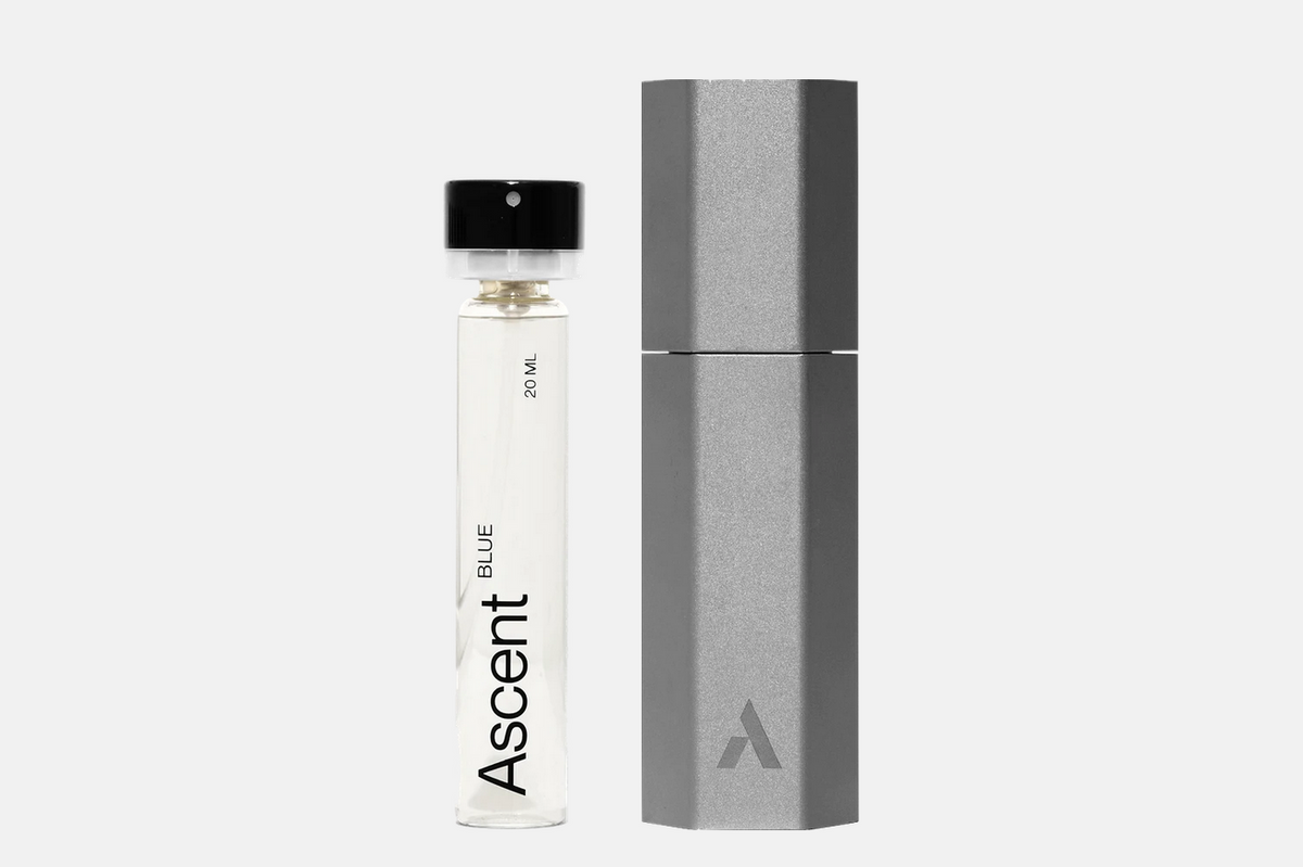 A cylindrical glass vial of Ascent Starter Pack - Blue perfume extract with a black cap, standing next to its hexagonal metallic carrying case.