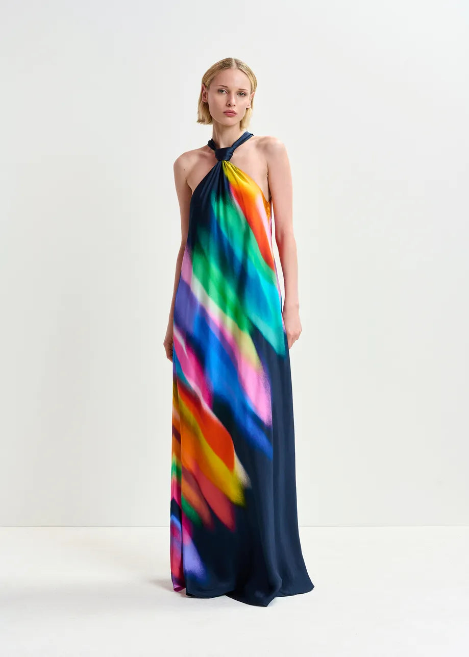 A person is wearing a vibrantly colored Essentiel Antwerp Halinki Halterneck Dress with a blurred pattern effect, standing against a plain white background.