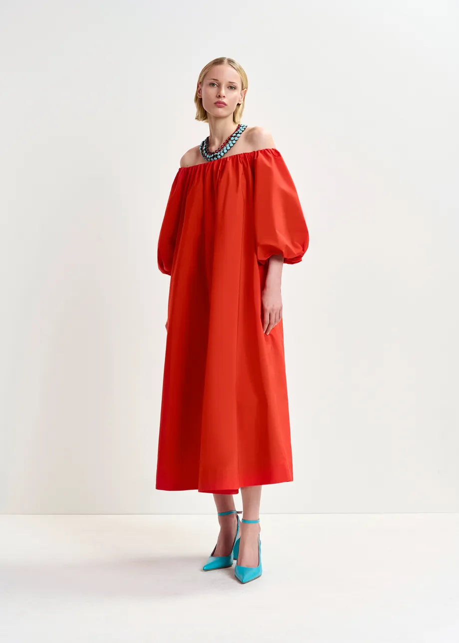 Wearing Essentiel Antwerp's Hamille Off-Shoulder Dress in red, featuring puffy sleeves and crafted from recycled polyester, a person poses elegantly against a minimal background, complemented by turquoise heels and a bold blue necklace.