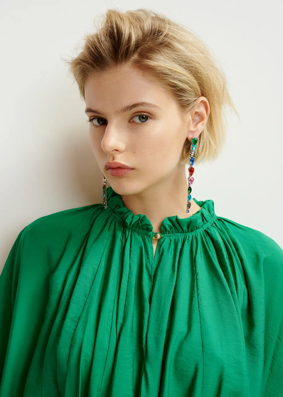 A person with short blonde hair wears Essentiel Antwerp's Silver Rhinestone Earrings - Multicolour and a vibrant green blouse, gazing towards the camera against a plain background.