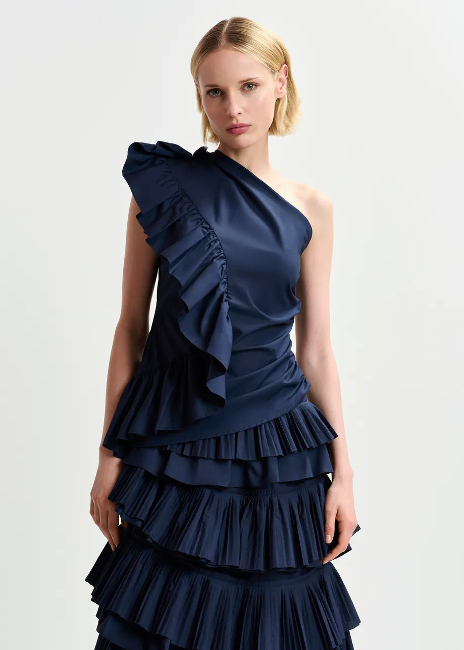 A person elegantly wears Essentiel Antwerp's Haven Top in dark blue, featuring a chic one-shoulder ruffle design, against a plain background.