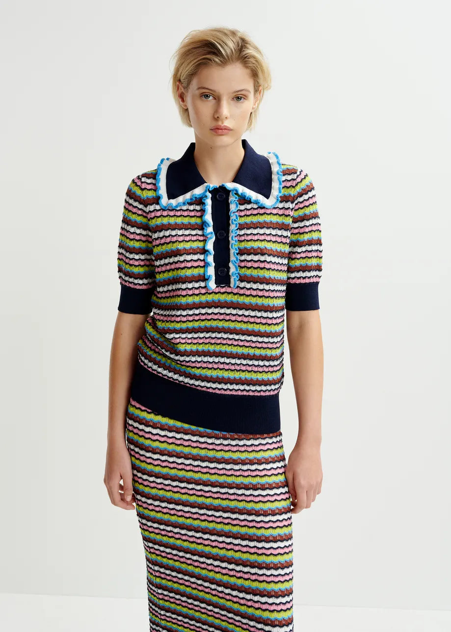 A woman stands against a plain background, wearing the Essentiel Antwerp Haximum Ruffled Polo in Multi with matching skirt, featuring a striped pattern and blue-trimmed collar for a stylish look.