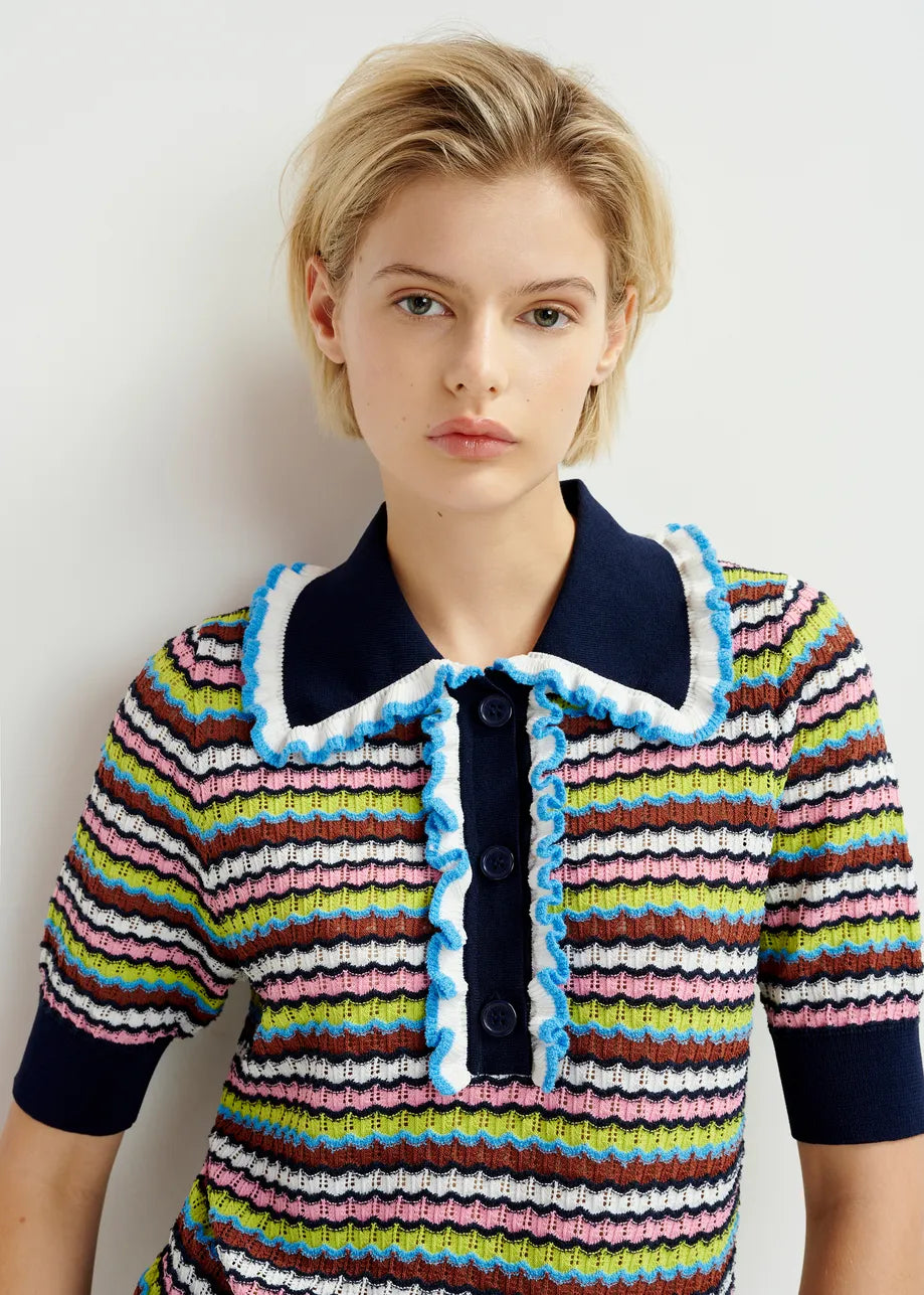 A person with short blonde hair models the Essentiel Antwerp Haximum Ruffled Polo - Multi, featuring colorful stripes, a navy collar, and blue-white ruffle trim, posed against a plain light background.