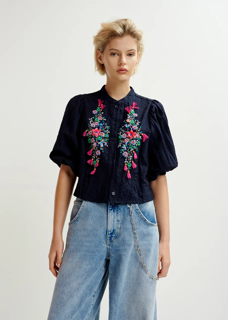 Someone is wearing the Heartfelt Shirt in navy from Essentiel Antwerp, featuring colorful floral sequin embroidery, paired with light blue jeans and a chic chain accessory.