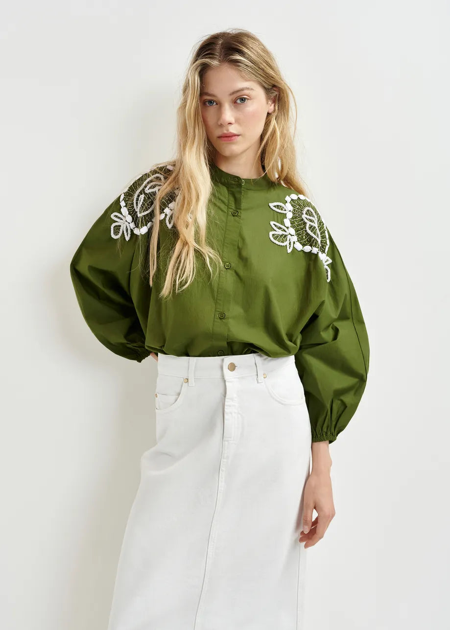 Someone is wearing the Hementine Shirt from Essentiel Antwerp, featuring blouson sleeves and intricate white floral embroidery on the shoulders, paired elegantly with a white skirt.