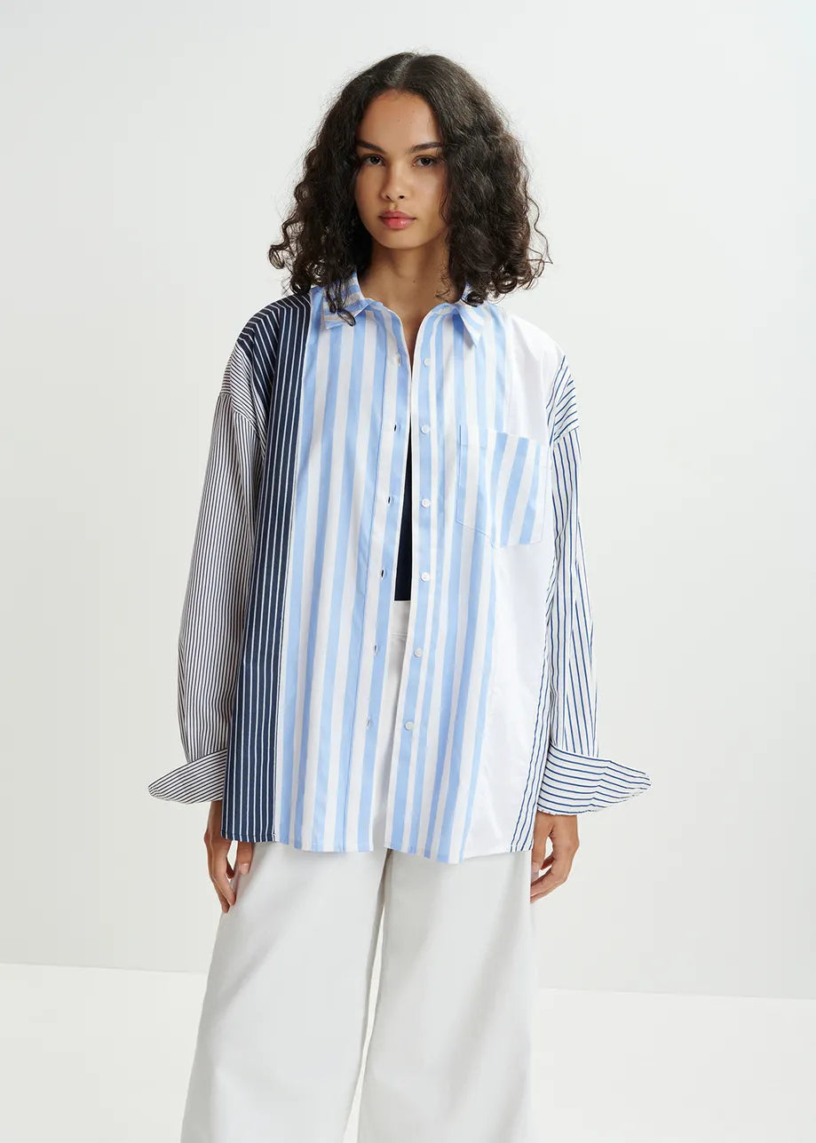 A woman exudes elegance in a loose-fitting oversized silhouette, wearing an Essentiel Antwerp's Hemix Striped Shirt - Blue. She pairs it effortlessly with wide white pants against a plain background.