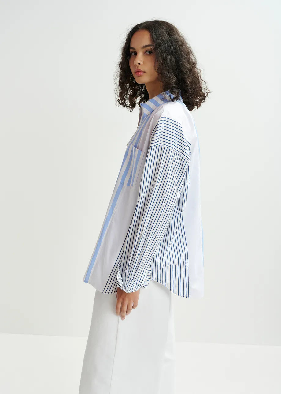 A person with curly hair stands in a minimalist setting, wearing an oversized outfit featuring Essentiel Antwerp's Hemix Striped Shirt in blue and white pants, exuding elegant style.