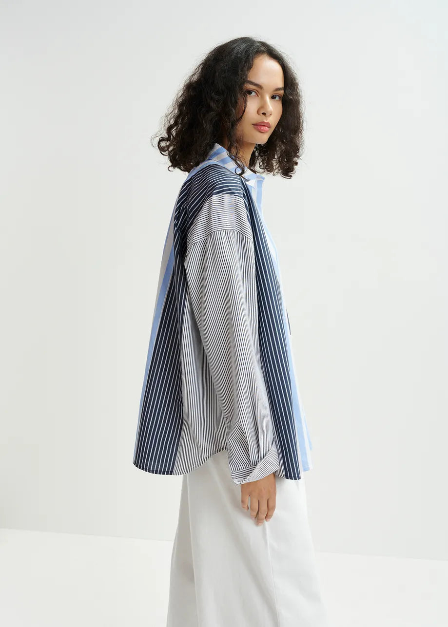 A person with curly hair in an Essentiel Antwerp Hemix Striped Shirt - Blue and white pants stands in a light room, exuding elegance while gazing sideways.