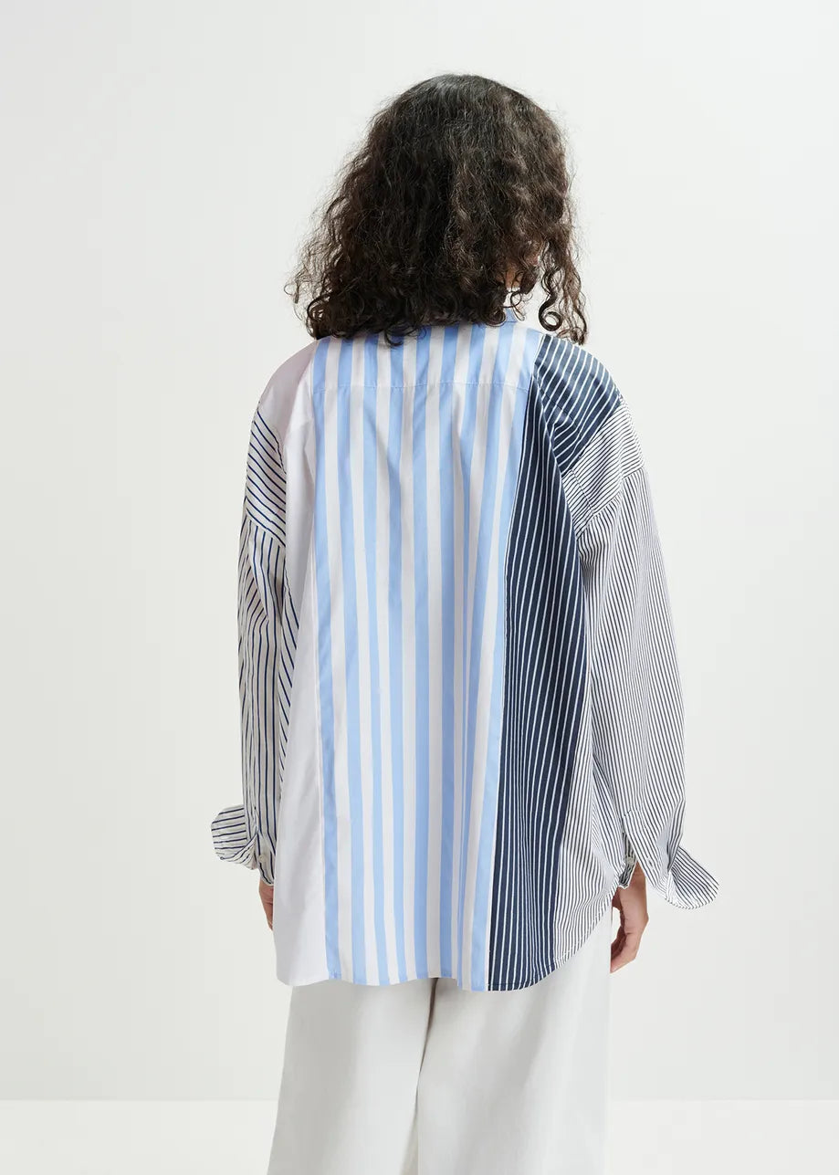 A curly-haired person wearing an oversized Hemix Striped Shirt in blue by Essentiel Antwerp, paired with white pants, stands against a plain background.