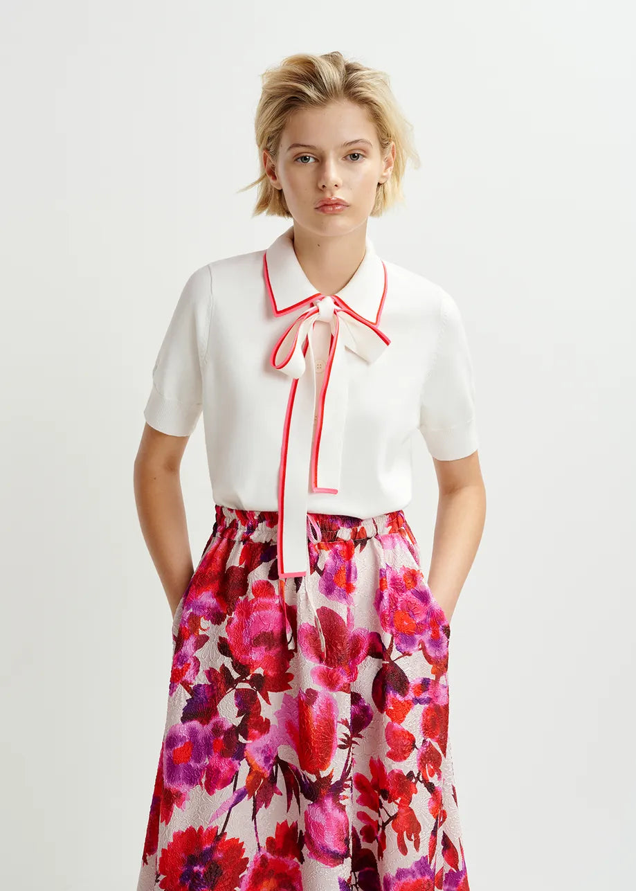 Wearing the Hight Knitted Polo top by Essentiel Antwerp, featuring chic ribbed trims and a red bow, a person stands against a plain background, pairing it beautifully with a floral skirt.